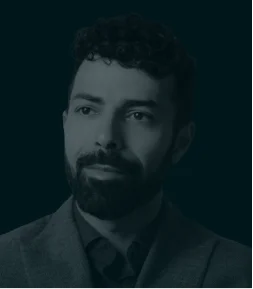 A portrait of Behrad Mirafshar with a short beard and curly hair, dressed in a suit jacket and shirt, gazing slightly to the side with a neutral expression. The image has a dark greenish tint, giving it a modern and professional tone